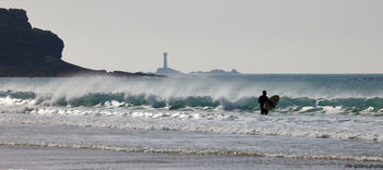Longships surfer This landscape photograph captures a surfer holding a surfboard, standing in the surf at Sennen Cove beach on the coast of Cornwall in the United Kingdom. Taken in the afternoon during the spring season, the image shows natural waves rolling toward the shore with a rugged rocky coastline on the left. In the distance, the Longships Lighthouse is clearly visible offshore, acting as a notable maritime landmark. The setting emphasizes the natural beauty of the area, with the bright spring sunlight illuminating the water and the beach, characteristic of the coastal environment around Sennen Cove in Cornwall.